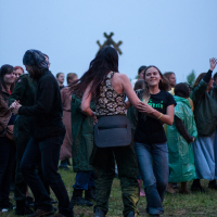 A lively outdoor gathering at dusk features a diverse group of people enjoying a dance at the 2012 Festival. The crowd, dressed in various casual and outdoor attire, smiles and chats. In the background, an indistinct sculpture adds to the festive atmosphere on the grassy area where some people are wearing raincoats.