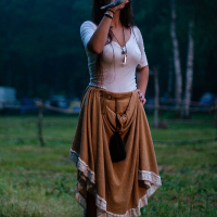 A person wearing a beige skirt and a white shirt stands outdoors in a grassy area, reminiscent of the serene settings at Kilkim Žaibu. The person is sipping a drink, with long hair and a small black pouch hanging from their waist. The backdrop reveals trees and a slightly blurred natural setting, suggesting a park or forest.