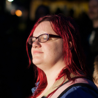 A person with long, bright red hair and glasses stands in a dimly lit area at Kilkim Žaibu 2012, looking to the right with a slight smile. They wear a blue jacket and an over-the-shoulder strap. The background is dark, with blurred lights and indistinct figures, creating a stark contrast with the well-lit subject.