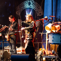 A band performs on stage with vibrant, colorful lighting at the 2012 Kilkim Žaibu festival. The group includes a female singer, a male guitarist, a male bagpipe player, and a male percussionist with drums. They are all dressed in medieval or fantasy-inspired costumes. Instruments and equipment are set up around them.