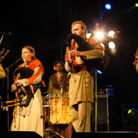 At the 2012 Kilkim Žaibu festival, a folk band performs onstage in medieval attire. Four musicians in beige robes and red hoods play bagpipes, while a drummer in the background plays a large drum. The stage is illuminated by soft yellow lights, with additional colorful lights enhancing the atmosphere.