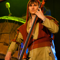 A person stands on stage wearing medieval-inspired clothing, including a green tunic and a red hooded cloak. They are playing an upright string instrument at Kilkim Žaibu 2012. Large rustic drums and drumsticks are visible in the background, and the stage is lit with yellow and blue lights.