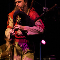 A person with long hair and a beard is playing a bagpipe on stage at the 2012 Kilkim Žaibu festival. They are dressed in beige and red medieval-style clothing. The background is dark, with faint purple and pink lights. Some musical equipment is visible in the background.