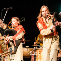 At the 2012 Kilkim Žaibu festival, a musical group performs on stage, featuring three members playing bagpipes and a drummer in the background. All members wear medieval-style costumes in beige and red. They are under stage lighting with a microphone stand visible in the foreground. The background is dark with some green stage lights.