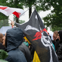People gather outdoors under cloudy skies at the 2012 Kilkim Žaibu festival. A person in a dark hooded jacket holds a pole with a large black flag featuring a white Jolly Roger with a red bandana, symbolizing a pirate theme. Another flag with red and white stripes is visible in the background, surrounded by green trees.