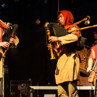 Three musicians, dressed in medieval-style tan robes with red hoods, perform on a dimly lit stage at the 2012 Kilkim Žaibu festival. Two play bagpipes, focused on their instruments, while the third in the background plays a string instrument. The scene evokes a historical or fantasy ambiance.