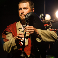 At Kilkim Žaibu 2012, a musician with short hair and a beard is playing a black bagpipe with a wooden pipe on stage. Clad in a medieval-style beige and red tunic, he performs under bright stage lights, which illuminate the dark background. A small camera is attached to his instrument.
