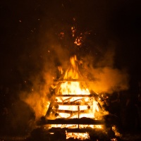 A large bonfire illuminates the night at Kilkim Žaibu 2012, with tall flames and sparks rising from a pyramid-shaped stack of logs. Dense smoke billows upwards, glowing orange in the firelight. The scene is set against a dark background, with shadows of onlookers faintly visible around the perimeter.