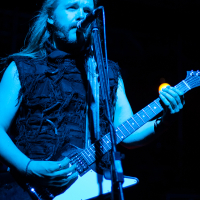 A guitarist with long hair performs passionately on stage under blue lighting, wearing a distressed, sleeveless shirt. He sings into a microphone while playing an electric guitar at Kilkim Žaibu 2012. The dark and intense atmosphere of the stage is enhanced by the focused lighting and shadows.