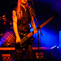 At Kilkim Žaibu 2012, a man with long hair performs on stage, playing a black electric bass guitar and singing into a microphone. He wears a sleeveless black top, revealing tattoos on his arms. Blue and purple stage lights create a dynamic atmosphere, while the drum kit with its logo is blurred in the background.