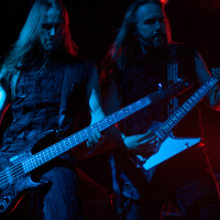 Two musicians perform energetically on stage at the 2012 Kilkim Žaibu festival under dramatic red and blue lighting. Both have long hair flowing and wear sleeveless black outfits. The musician on the left plays a bass guitar, while the one on the right plays a guitar. Amplifiers are visible in the background.