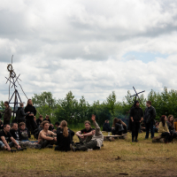 A group of people dressed in medieval attire sit and stand on a grassy field under a cloudy sky during Kilkim Žaibu 2012. Some engage in conversation, while others gaze around. Wooden structures with wheel-like designs stand in the background, and a treeline is visible further back.