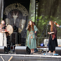 A folk music band performs on an outdoor stage at Kilkim Žaibu 2012. The group consists of five members: one playing a drum, another with maracas, a third with a guitar, and two singing into microphones. They are dressed in traditional, earthy-toned clothing. A banner with the band's name and a deer skull symbol is in the background.