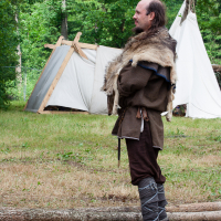 A person dressed in medieval-style clothing stands in a grassy area, reminiscent of Kilkim Žaibu 2012. The outfit includes fur over the shoulders, a brown tunic, and wrapped boots. Two white canvas tents framed by wooden poles are in the background amidst a lush, forested area. Logs lie on the ground near the person.