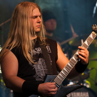 A musician with long blonde hair, wearing a black sleeveless shirt with a graphic design, plays an electric guitar on stage at the 2012 Kilkim Žaibu festival. He has a serious expression, and the background features a drummer, drums, and a "Yamaha" bass drum. The stage is dimly lit with blue and green lighting effects.