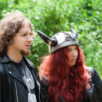 A young man and woman, both with long wavy hair, stand side by side outdoors, looking like they're ready for Kilkim Žaibu. The man has light brown hair, a short beard, and is wearing a black leather jacket. The woman has bright red hair flowing down her shoulders beneath a horned helmet. Trees and greenery fill the background. It's 2012 at the music festival