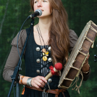 A woman with long brown hair stands on stage at the Kilkim Žaibu Festival, wearing a black dress with silver buttons and a brown shirt underneath. She is holding a handheld drum in her left hand and a mallet in her right, singing into a microphone in front of a green background. She wears a narrow headband and several necklaces.