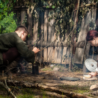 Two individuals, dressed in historical attire and enveloped by forest greenery, engage in activities outside a rustic wooden hut with a thatched roof. One person kneels to tend to a small fire with a stick while the other prepares food at what seems like an early 2012 Kilkim Žaibu reenactment amidst various cooking supplies.