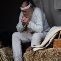 A man dressed in a white medieval-style tunic and headband sits on a bundle of hay at the Kilkim Žaibu festival, 2012. In the dimly lit setting, he appears to be focusing intently on something he is holding in his hands. An empty brown basket is placed on a nearby hay bundle.