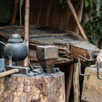 A blacksmith's workspace featuring a weathered anvil, various hammers, and tools scattered on a tree stump. Nearby is a teapot with a handle and spout. The background displays wooden structures and green foliage, reminiscent of the rustic ambiance at Kilkim Žaibu 2012.