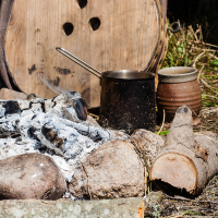 A campfire site with charred logs and white ashes is surrounded by a circle of large rocks, reminiscent of a Kilkim Žaibu festival in 2012. In the foreground, there are logs and firewood. A metal pot and ceramic mug rest near the shelter wall, constructed from wooden planks with three small holes. Grassy ground is visible around.