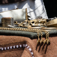 Close-up of a table covered with a brown cloth at the 2012 Kilkim Žaibu festival, displaying various handmade brass and bronze jewelry and accessories, including bracelets, rings, and necklaces. The items showcase intricate designs and craftsmanship. Sunlight casts soft shadows, highlighting the metallic texture of the pieces.