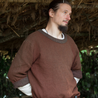 A person with long dark hair tied back and a light beard is standing under a thatched roof at the 2012 Kilkim Žaibu festival. They are wearing a brown tunic over a white shirt and have a belt with a leather pouch and a sheathed knife at their waist. They are looking to the right with their hands clasped behind their back.
