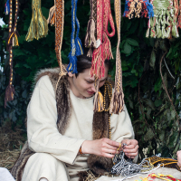 At Kilkim Žaibu festival in 2012, a person is seated and working with textiles, surrounded by many colorful, intricately patterned woven bands hanging above. They are dressed in light-colored, historical-looking clothing with fur accents and are focused on their task, situated in a natural setting with greenery and hay.