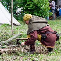 A person dressed in medieval clothing, including a brown tunic, olive green headscarf, and cloak, crouches down to chop wood with an axe. The scene takes place in an outdoor camp during the 2012 Kilkim Žaibu festival, with greenery and a canvas tent. In the background, people are gathered, partially obscured by the foliage.