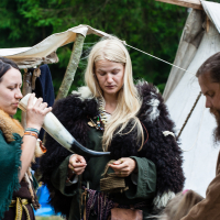 Three people are dressed in medieval or Viking-style clothing in front of a canvas tent at Kilkim Žaibu 2012. The person on the left drinks from a large horn, the one in the middle looks down while holding an object, and the person on the right has their head bowed, perhaps focusing on something. Trees are visible in the background.