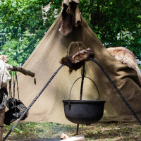 A 2012-inspired camping setup features a canvas tent nestled among green foliage. In front of the tent, a black kettle hangs from a tripod over a smoldering fire pit with logs. Fur pelts are draped over the tent, and a wooden branch serves as a makeshift hanger for clothes and gear, reminiscent of Kilkim Žaibu festival vibes.