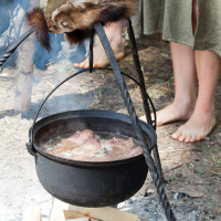 A black cast iron pot hangs over an open fire, suspended by a tripod made of twisted metal rods. The pot contains simmering food. A fur-lined hat rests on top of the tripod. Barefoot individuals wearing historical garments stand on the grass in the background, capturing a moment reminiscent of Kilkim Žaibu 2012.