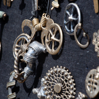 Close-up image of various steampunk-style metal trinkets and gears attached to a black textured background. The items include cogs, keys, wheels, and intricate mechanical parts, showcasing a blend of industrial and Victorian aesthetics reminiscent of the Kilkim Žaibu 2012 festival. The metallic pieces vary in color, primarily bronze and silver.