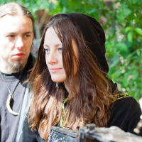 A young woman with long brown hair, wearing a black hood, smiles softly while looking to her right. A man with a goatee beard and long brown hair stands behind her, gazing in the same direction. They are outdoors at Kilkim Žaibu 2012, surrounded by lush greenery. Both are wearing black clothing and necklaces.