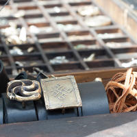 A close-up image of a display featuring handcrafted metal bracelets and leather accessories laid out on a wooden surface at Kilkim Žaibu 2012. The foreground showcases two distinct metal bracelets with intricate designs, one circular and one square, alongside coiled leather strips. Blurred background reveals more jewelry.