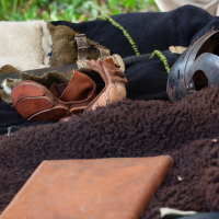 A scene of medieval camping items: a leather-bound journal rests on a dark fur blanket, surrounded by brown leather gloves and a metal helmet. The background includes various fabric and leather goods, suggesting a historical reenactment setting, reminiscent of Kilkim Žaibu 2012. Grass is visible in the distance.