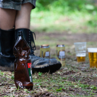 Close-up of a person wearing black rain boots and shorts standing on a forest floor with scattered leaves and grass at the 2012 Kilkim Žaibu festival. In the foreground, a crumpled brown plastic bottle lies on the ground. In the background, several beer cans and plastic cups filled with beer are scattered about.