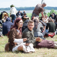 A group of people, some in medieval costumes, are gathered on a grassy field by a body of water at the 2012 Kilkim Žaibu festival. A seated pair in the foreground, one with red hair and the other a blond man, are holding bowls. In the background, others are sitting and interacting, with trees and water visible beyond.