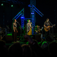 A band performs on a dark stage featuring colorful lights and rigging at the 2015 Kilkim Žaibu festival. The group comprises five members: three women, one man with a guitar, and another man with a keyboard. Audience members can be seen in the foreground, some with raised hands. The backdrop includes various musical equipment.