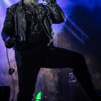 A person in a black leather outfit and dramatic face paint passionately sings into a microphone on a dimly lit stage at the Kilkim Žaibu festival. Blue and purple lights illuminate the background, musical equipment including speakers and guitars are visible, and smoke adds a mysterious atmosphere, enhancing the performance mood.