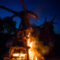 At the Kilkim Žaibu festival in 2015, a tall, human-like sculpture made of straw is engulfed in flames at night, set against a dark blue sky. The burning straw emits bright orange and yellow flames, creating a stark contrast with the surrounding darkness. Another similar but unlit sculpture is visible in the background.