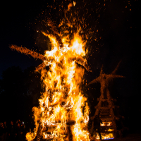 During the 2015 Kilkim Žaibu festival, a large straw effigy is engulfed in bright, intense flames at nighttime, sending sparks and embers into the dark sky. A crowd of spectators watches the bonfire from a distance. Another smaller burning effigy is visible in the background. The ground is covered in straw.