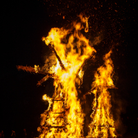 Two large wooden structures engulfed in bright, intense flames during the night at the 2015 Kilkim Žaibu music festival. The fire illuminates the dark surroundings, casting a dramatic glow. A small crowd of silhouetted figures can be seen in the distance observing the burning structures.