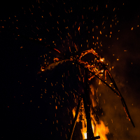 A large fire burns in the dark, with bright orange and yellow flames illuminating a skeletal structure within the blaze. Sparks fly upward, creating a dramatic and intense scene. The image captures the dynamic and destructive nature of the fire against a pitch-black background at Kilkim Žaibu 2015 music festival.