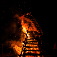 A large wooden structure burns intensely in the night at the 2015 Kilkim Žaibu music festival, emitting bright flames and sparks. The dark sky and surrounding area are illuminated by the fire, with silhouettes of people visible in the background. The structure appears to be a tall, slatted frame built from logs and branches.
