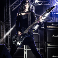 A musician with long black hair, wearing a black outfit, plays a bass guitar passionately on stage at the 2015 Kilkim Žaibu festival. Surrounded by metal scaffolding and numerous Marshall amplifiers, the stage is lit with strong white and blue lights, while water bottles lie on the floor in the foreground.