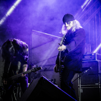Two musicians perform on a dimly lit stage with blue and purple spotlights at Kilkim Žaibu 2015. The guitarist on the right wears a beanie and dark coat, playing intently, while the bassist on the left, with long hair covering their face, is deeply engrossed in playing. Amplifiers and stage lighting are visible in the background.
