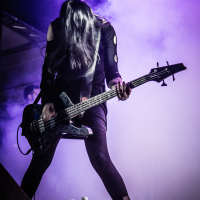 At the 2015 Kilkim Žaibu festival, a woman wearing a black outfit passionately plays a bass guitar on stage, surrounded by a purple mist. Her long hair obscures her face as she leans forward, immersed in her performance. Two water bottles and effects pedals are visible on the ground in front of her. Lighting highlights her dynamic presence.