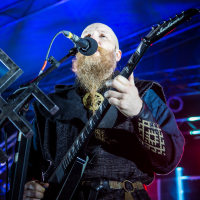 A musician with a long, curly beard plays an electric guitar on stage at the 2015 Kilkim Žaibu festival. He is singing into a microphone and dressed in a dark, medieval-inspired outfit. The background is lit with blue and red stage lights, creating an atmosphere that suggests a live concert performance.