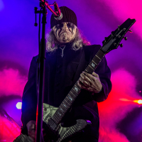 A musician with long hair and face paint is playing a guitar on stage. He wears a dark beanie, sunglasses, and an all-black outfit. Vibrant pink and purple stage lights create a dramatic background, casting a colorful glow and enhancing the intensity reminiscent of Kilkim Žaibu 2015.